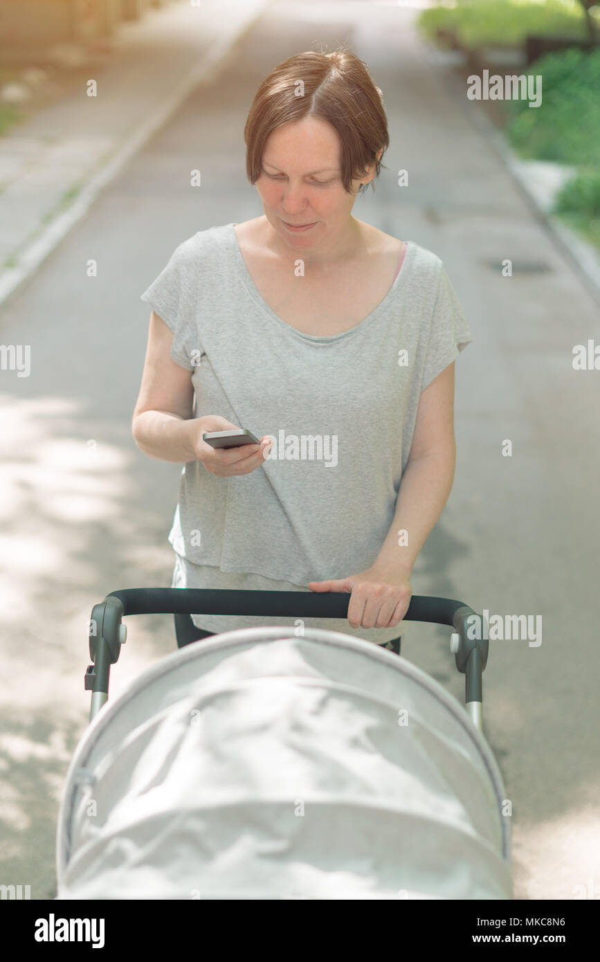 Mère poussant poussette de bébé et la lecture de texte message sur smart phone en marchant sur la rue en vêtements de sport décontracté Banque D'Images