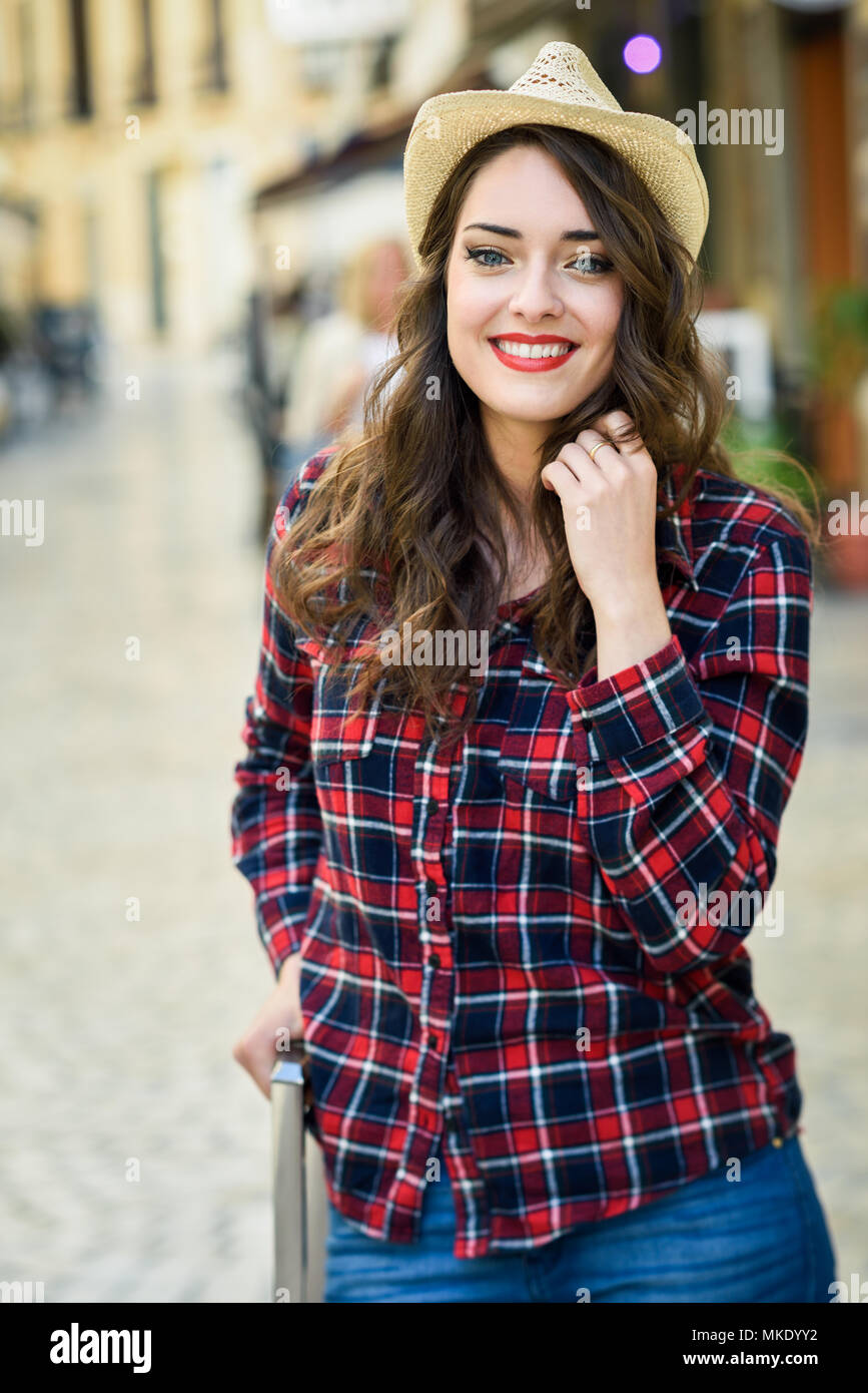 Jeune femme avec de beaux yeux bleus portant chemise à carreaux et chapeau de soleil. Girl smiling in contexte urbain. Banque D'Images