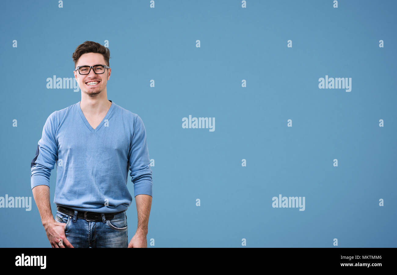 Jeune homme séduisant en chandail et lunettes à la caméra en se tenant debout sur fond bleu. Banque D'Images