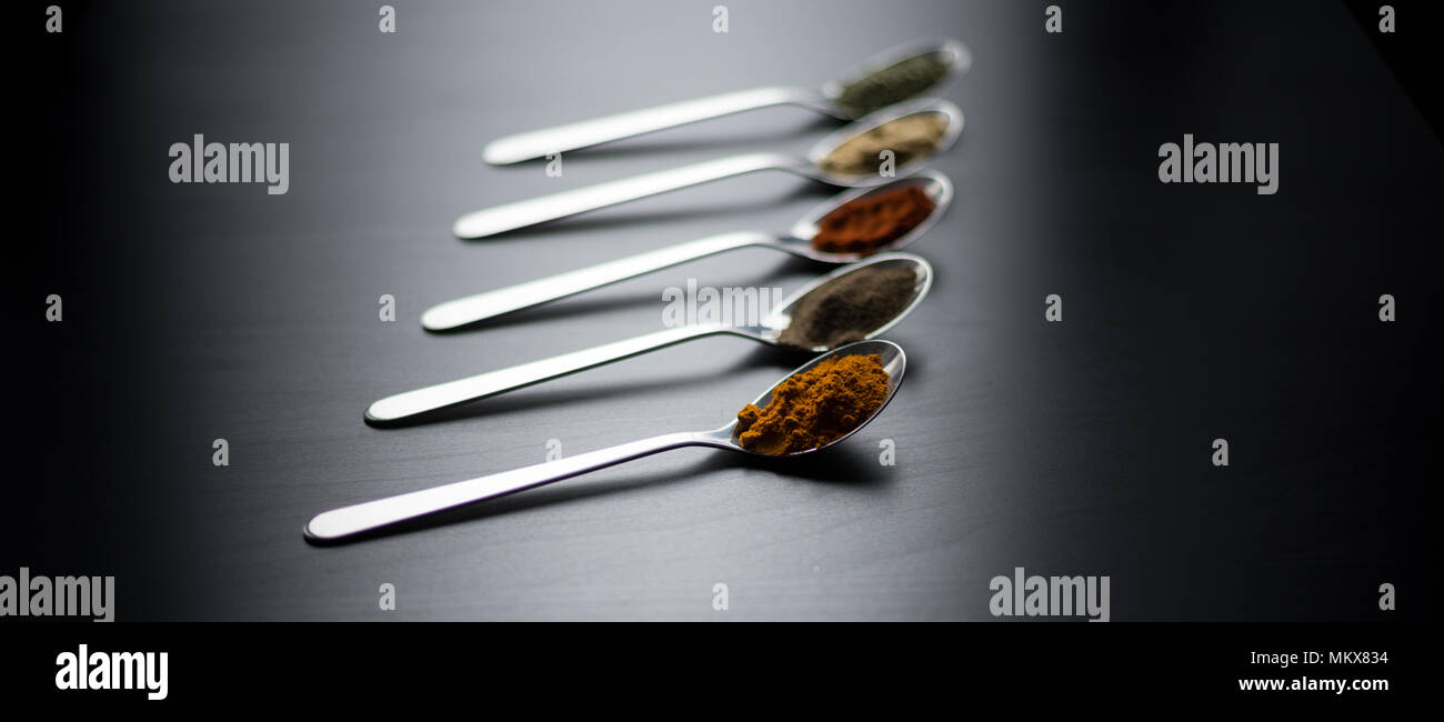 Épices colorés en cuillères d'argent, posé sur une table en bois noir. Poivre, sel, poivre, basilic, moutarde Banque D'Images