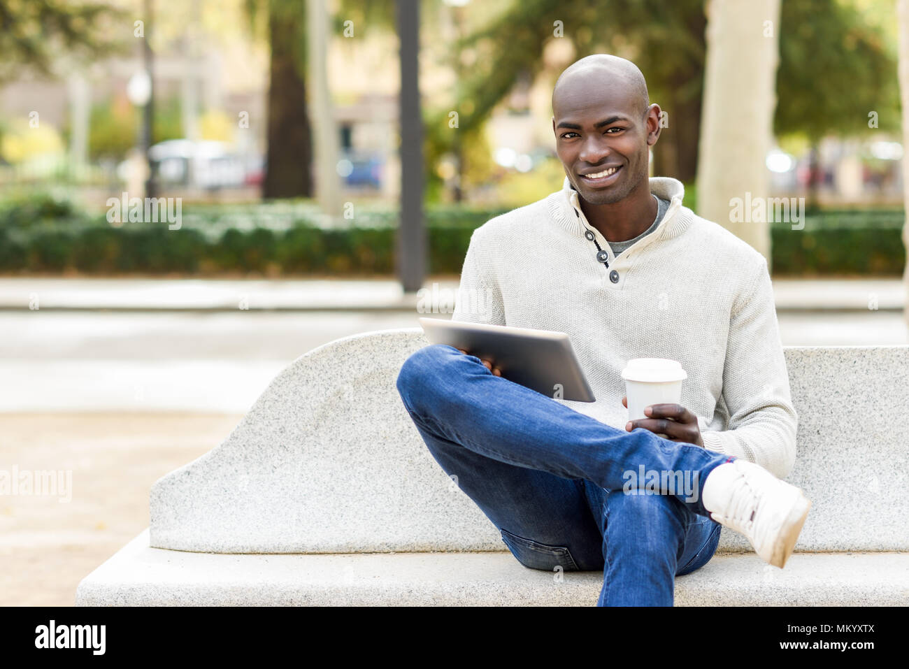 Black tablet et café à emporter en contexte urbain. Jeune africain guy avec crâne rasé portant des vêtements décontractés. Banque D'Images