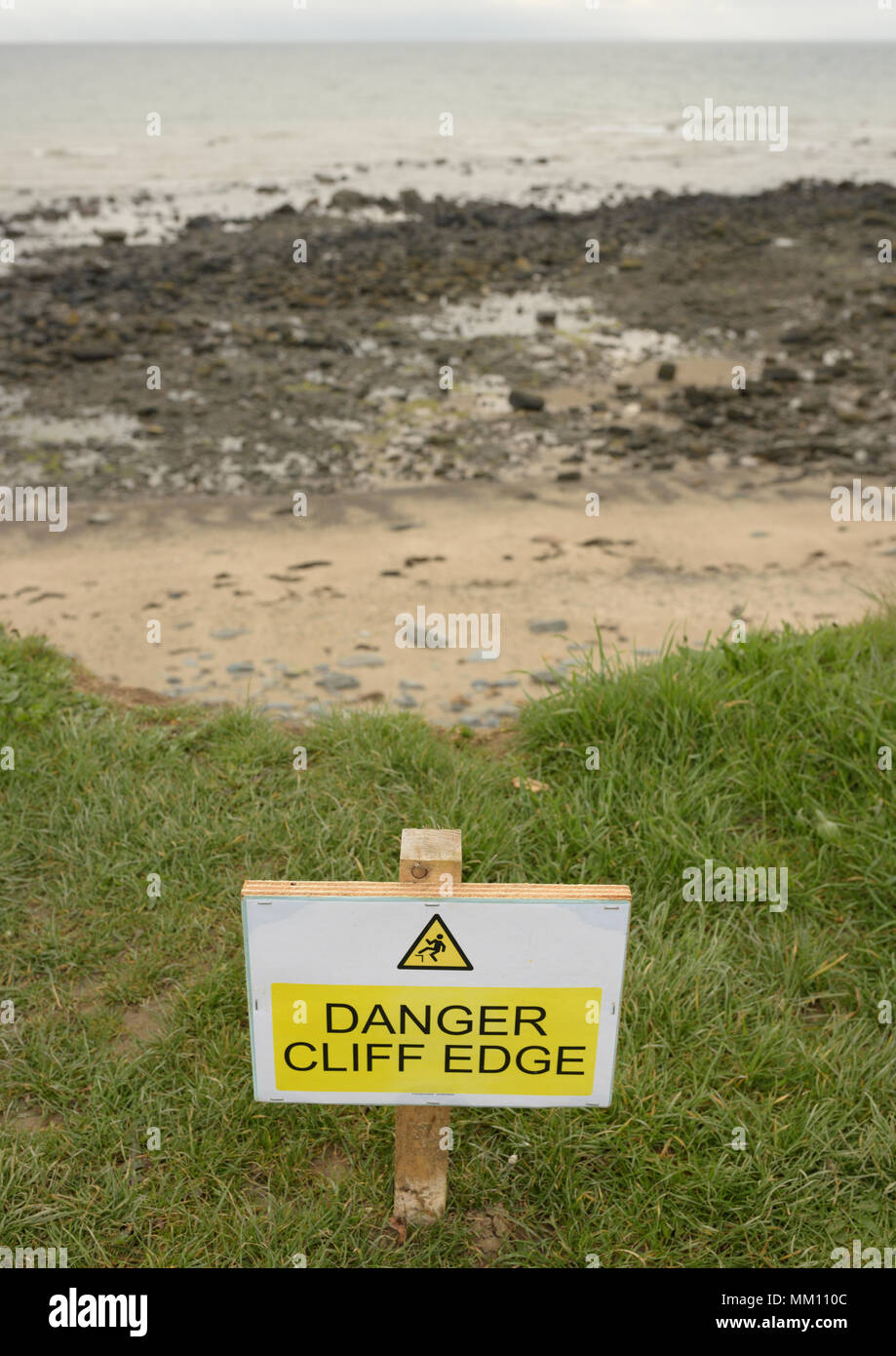 Panneau d'avertissement jaune et blanc de bord de falaise danger sur le chemin de la côte du pays de galles, plage, marée basse en arrière-plan, érosion côtière près de Pwllheli côte nord du pays de galles uk Banque D'Images