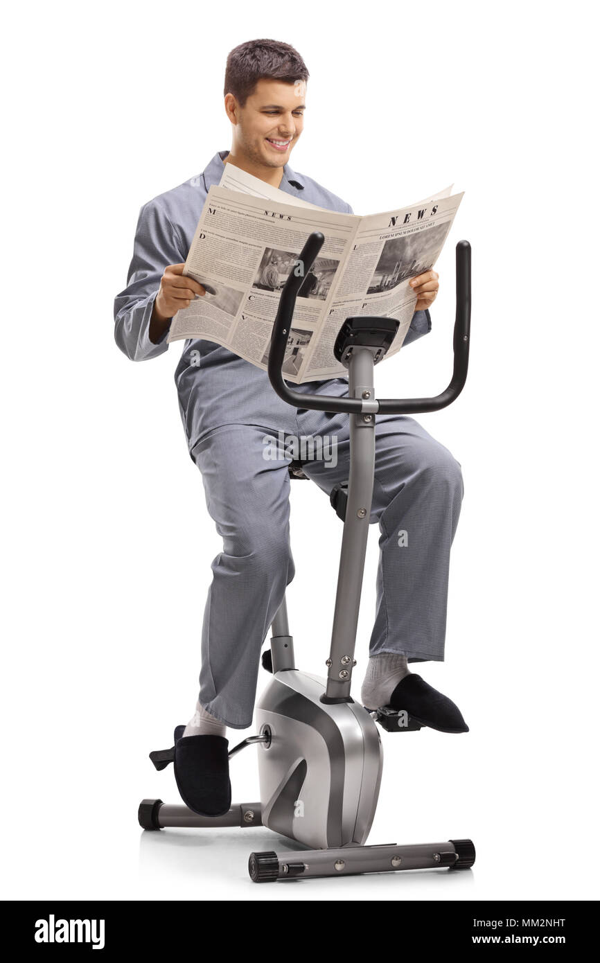 Jeune homme en pyjama lire un journal sur un vélo stationnaire isolé sur fond blanc Banque D'Images