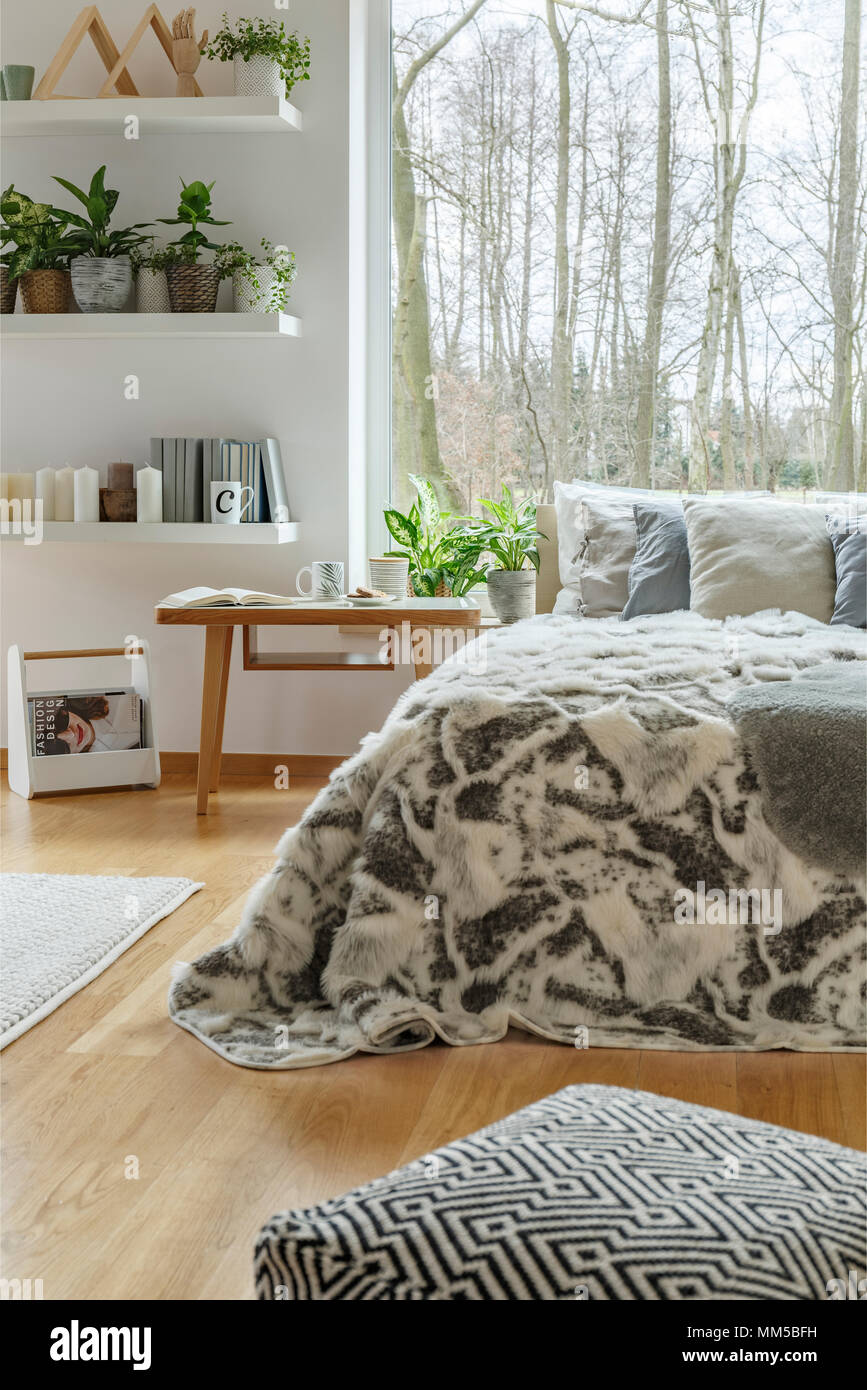 Pouf à motifs près de lit avec la fourrure dans une chambre confortable avec des plantes d'intérieur et vue sur une forêt Banque D'Images