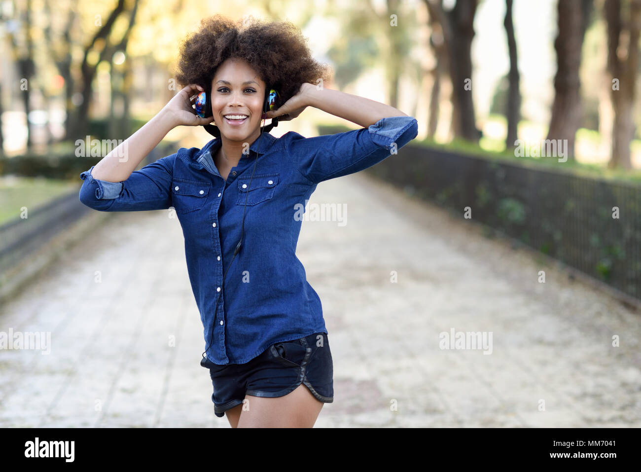 Jeune femme noire avec la coiffure afro à écouter la musique avec les écouteurs en contexte urbain. Drôle mixte woman wearing blue shirt et shorts. Banque D'Images