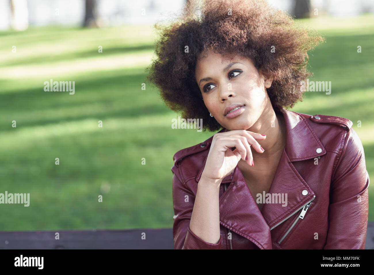 Jeune femme noire avec afro hairstyle assis dans un banc dans un parc urbain. Woman wearing red mixte cuir veste et robe blanche avec city backgroun Banque D'Images