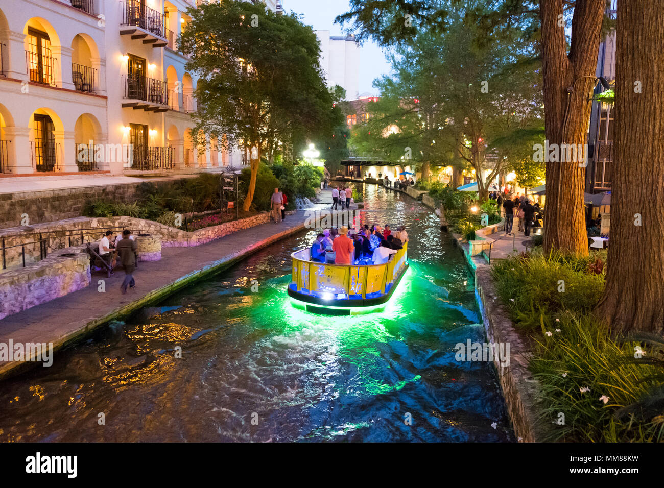 San Antonio, Texas - 18 Avril 2018 : bateaux traversent le fleuve avec des piétons sur les trottoirs de la promenade le long de la rivière dans le centre-ville de San Antonio à twilig Banque D'Images
