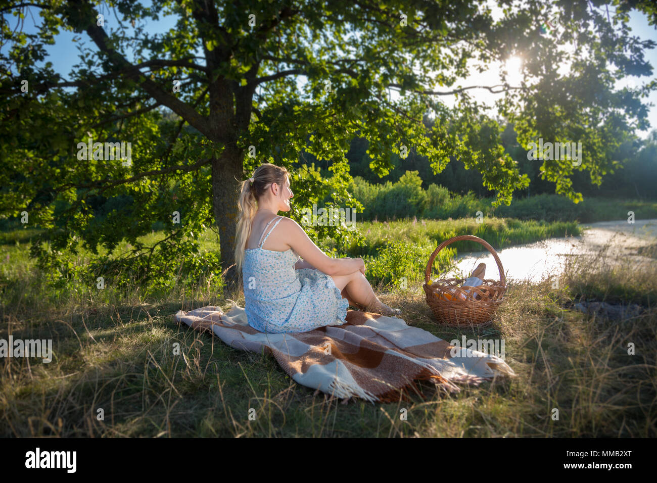 Belle jeune femme assise sur une couverture sous grand arbre près du lac et à la recherche au coucher du soleil Banque D'Images