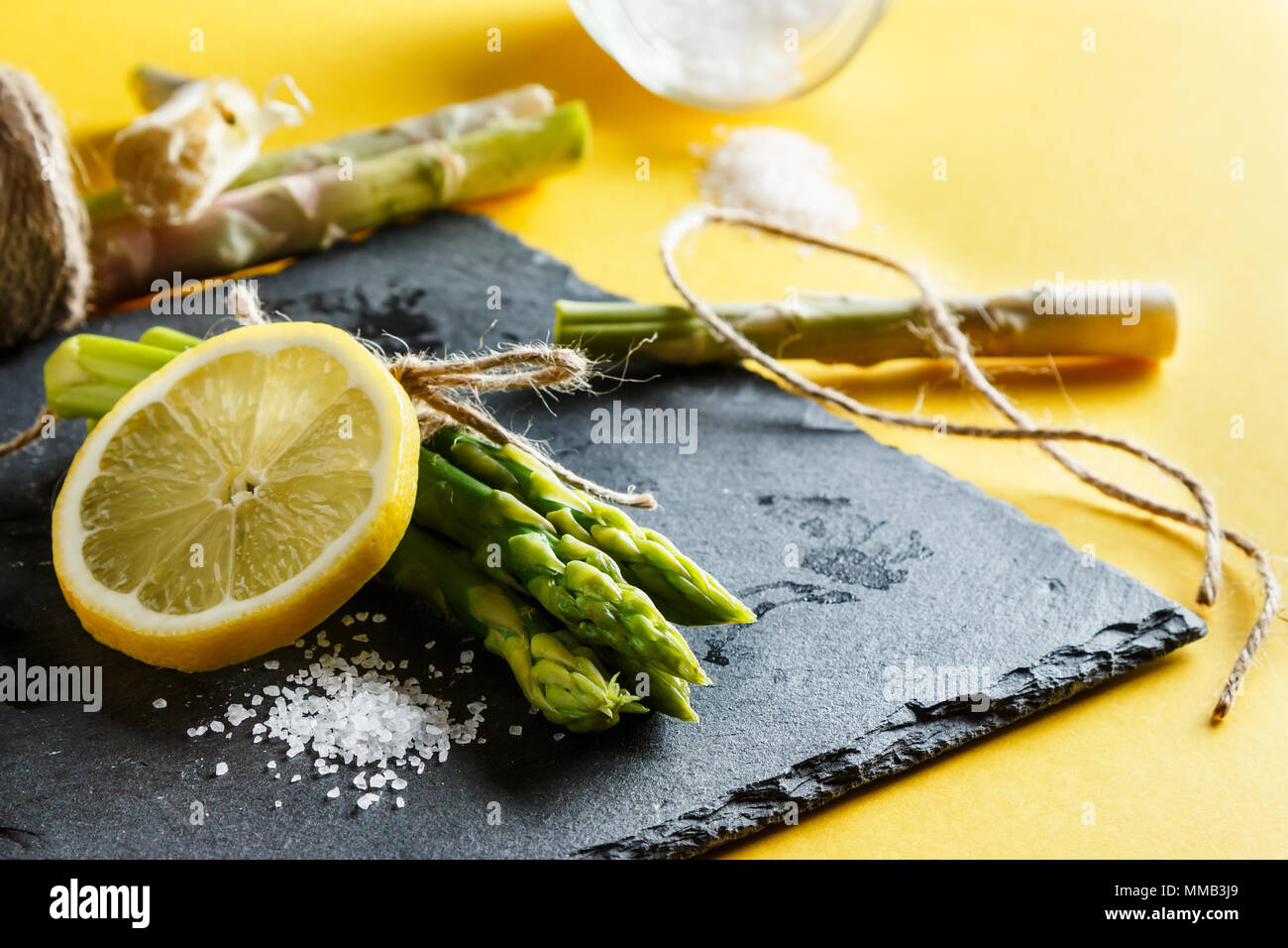 Les asperges. Bunch of fresh green asparagus fermer jusqu'au gros sel et d'une tranche de citron sur une plaque en ardoise sur fond jaune. Morceaux de corde et esparr Banque D'Images