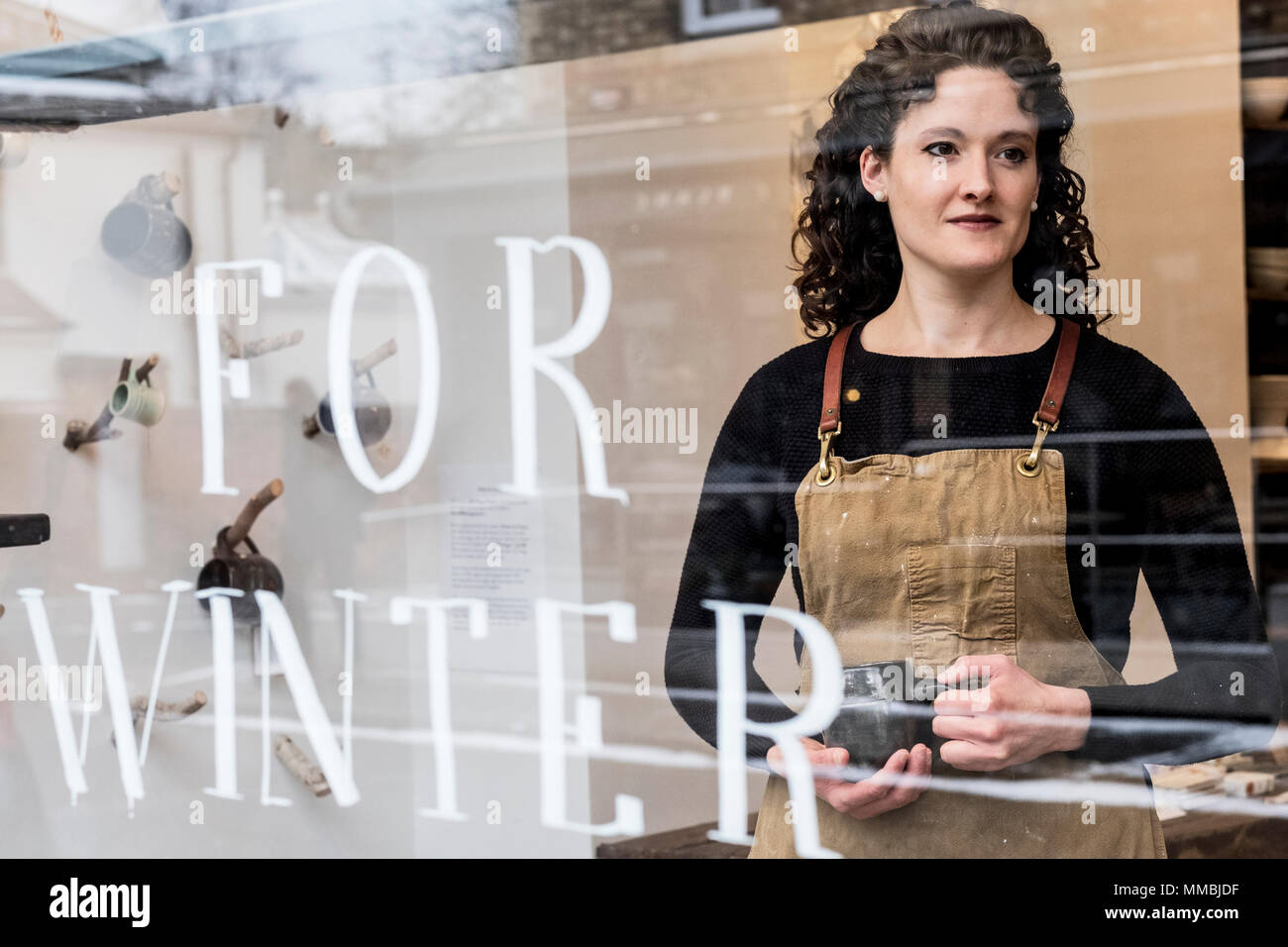 Femme aux cheveux bouclés brun Portant tablier, debout derrière des fenêtres, magasin de poterie holding mug. Banque D'Images