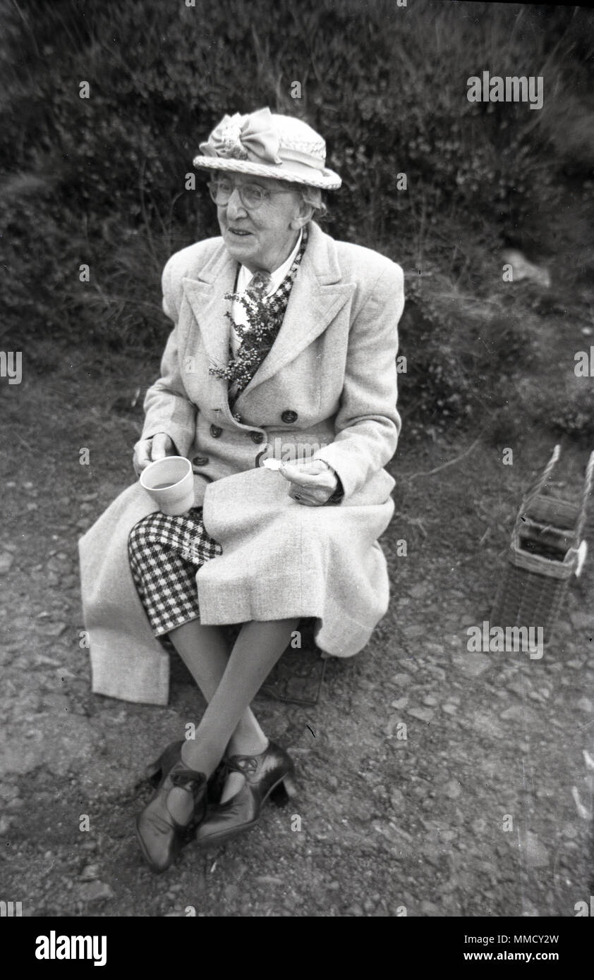 Années 50, une vieille dame bien habillé portant un chapeau et enduire  assis sur un chemin à l'extérieur, avoir une tasse de thé et une collation,  England, UK Photo Stock - Alamy