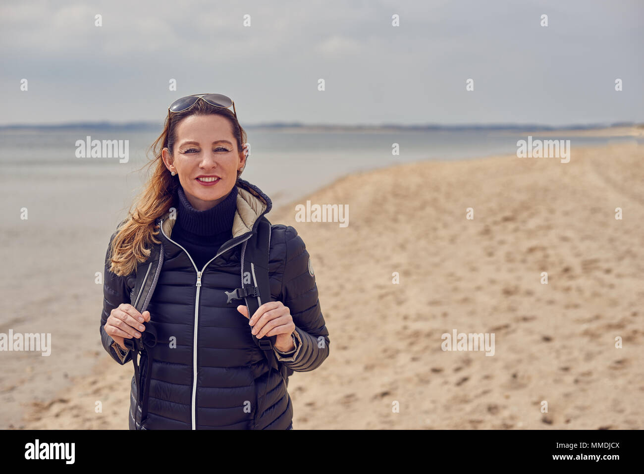 Jolie femme en bonne santé bénéficiant d'une randonnée sur la plage un jour nuageux froid debout souriant à la caméra sur le sable tenant les sangles de son retour Banque D'Images