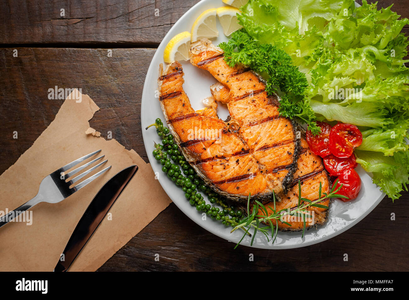 Pavé de saumon grillé servi avec des tomates grillées, salade, tranches de citron, graines de poivre, romarin, persil sur plaque blanche sur table en bois. Banque D'Images