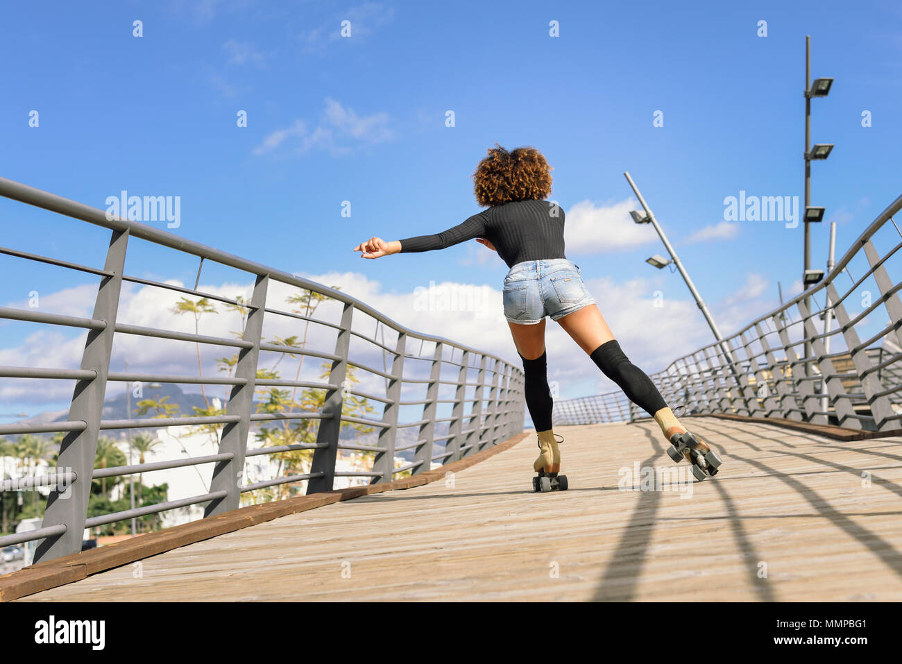 Vue arrière de femme noire, coiffure afro, équitation sur patins à l'extérieur sur le pont urbain. Jeune fille à roues alignées sur un jour ensoleillé. Banque D'Images