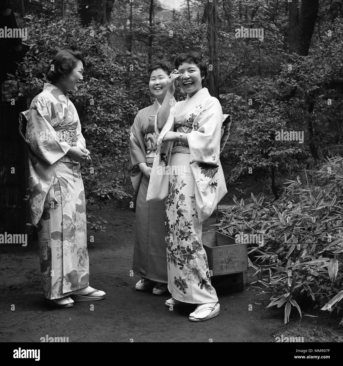 Années 1950, tableau historique, trois jeunes femmes se tenant ensemble à l'extérieur dans un jardin japonais de porter le costume national, le kimono, un chiffon slik enroulé autour du corps, liée à l'il taille, avec une obi, une large ceinture. Banque D'Images