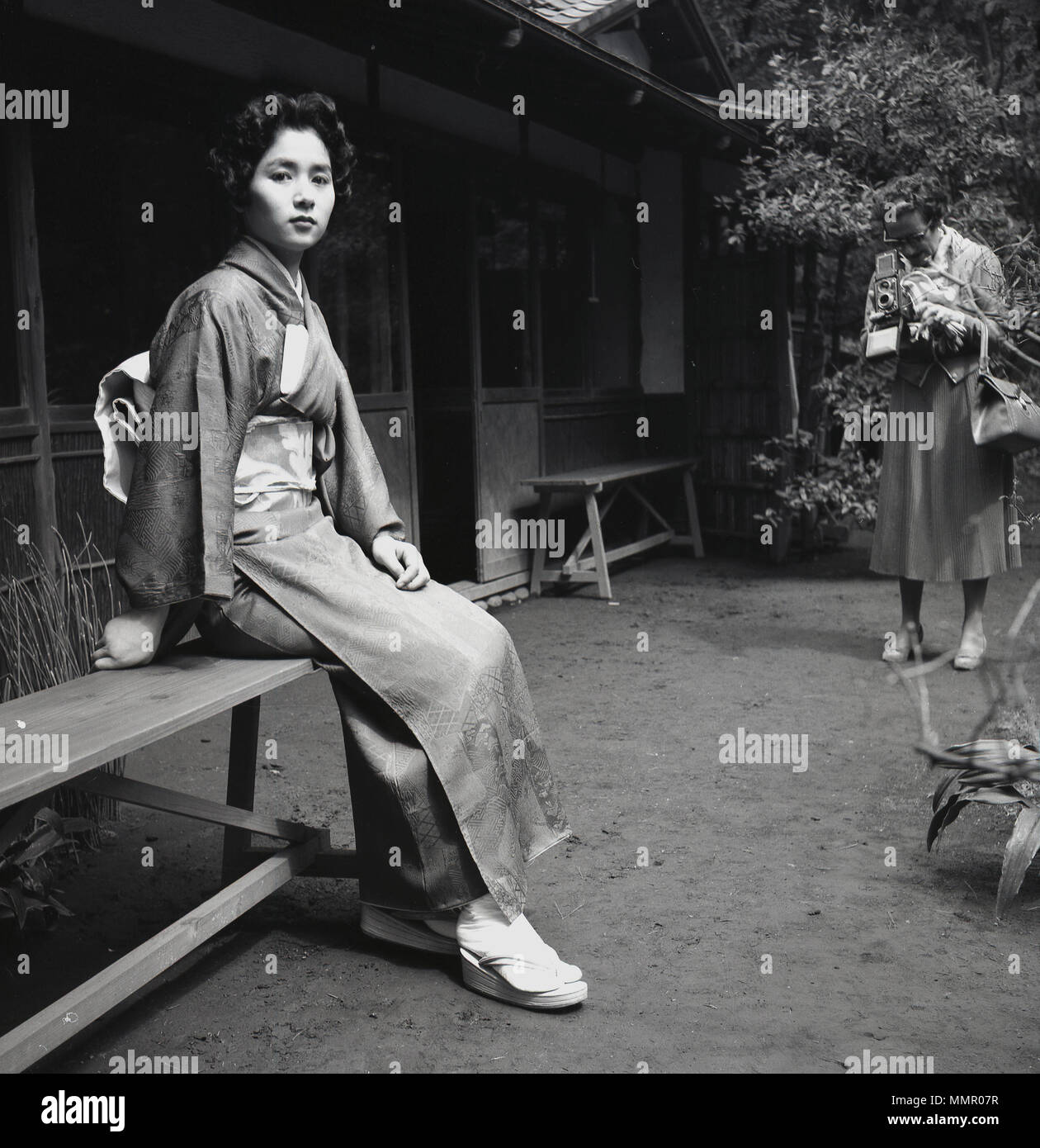 Années 1950, historiques, séduisant jeune femelle adulte assis sur un banc à l'extérieur portant les vêtements traditionnels japonais ou costume, le kimono et photographié par une dame de l'ouest. Le kimono est un vêtement de soie qui est enroulée autour du corps, attachés avec une large ceinture, un obi et est la tenue nationale pour le Japon. Banque D'Images