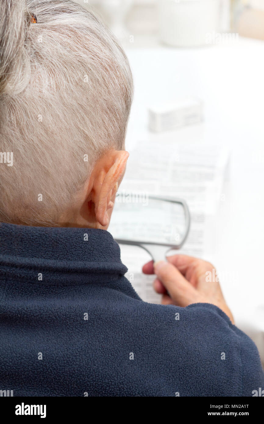Vieille Femme assise à une table dans son salon à l'aide d'une loupe pour lire la feuille d'information de ses médicaments prescrits Banque D'Images