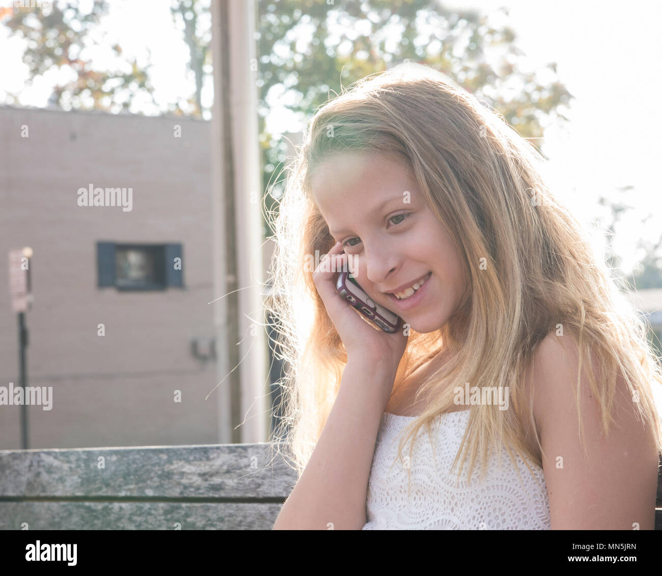 10 ans, fille, assise sur un banc à parler sur son téléphone cellulaire, en souriant. Banque D'Images