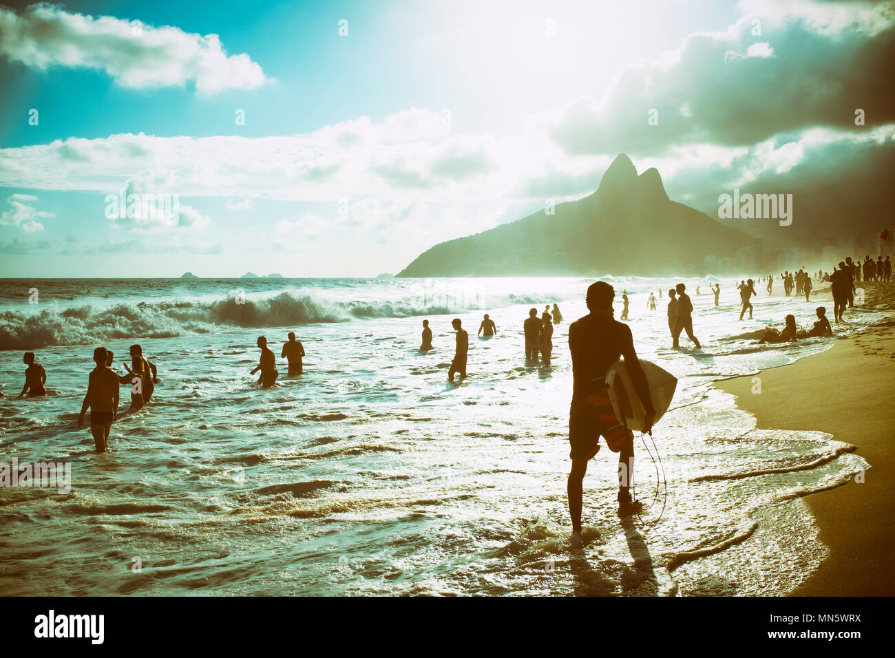 Silhouette d'un surfeur qui sort des vagues avec sa planche de surf en face d'une vue sur le coucher de soleil de deux frères montagne sur la rive de la plage d'Ipanema Banque D'Images
