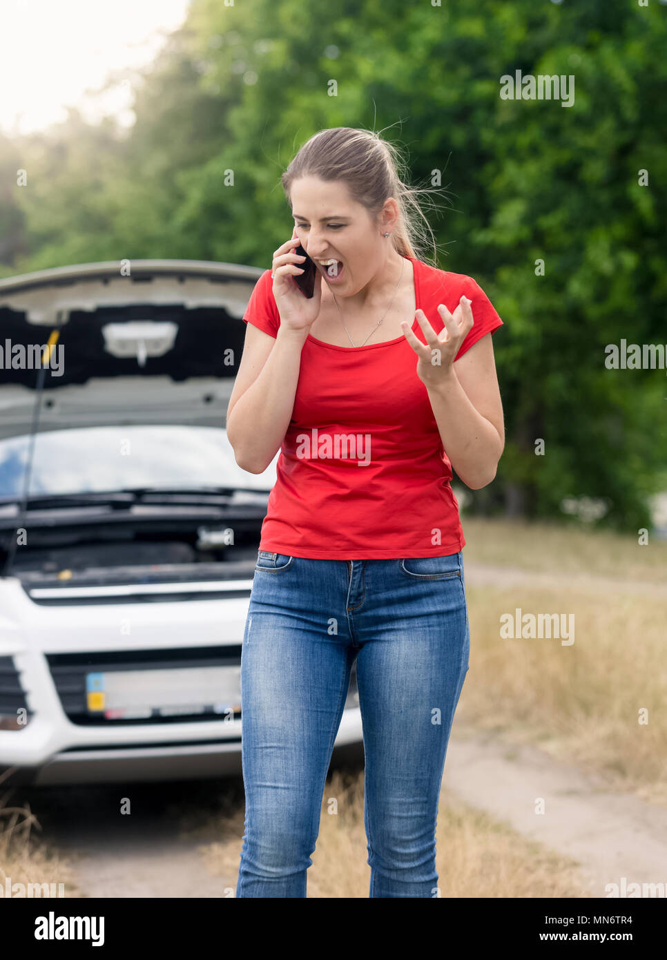 Angry young woman standing at voiture cassée dans le champ et de crier au téléphone mobile Banque D'Images