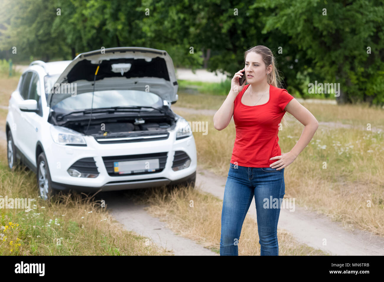 Jeune femme en colère debout à sa voiture cassée dans terrain et parler par téléphone avec le mécanicien Banque D'Images