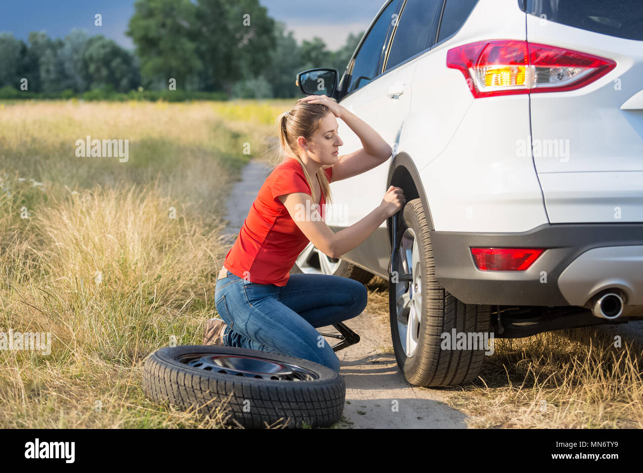 Jeune femme luttant pour changer un pneu de voiture à plat sur route de campagne Banque D'Images