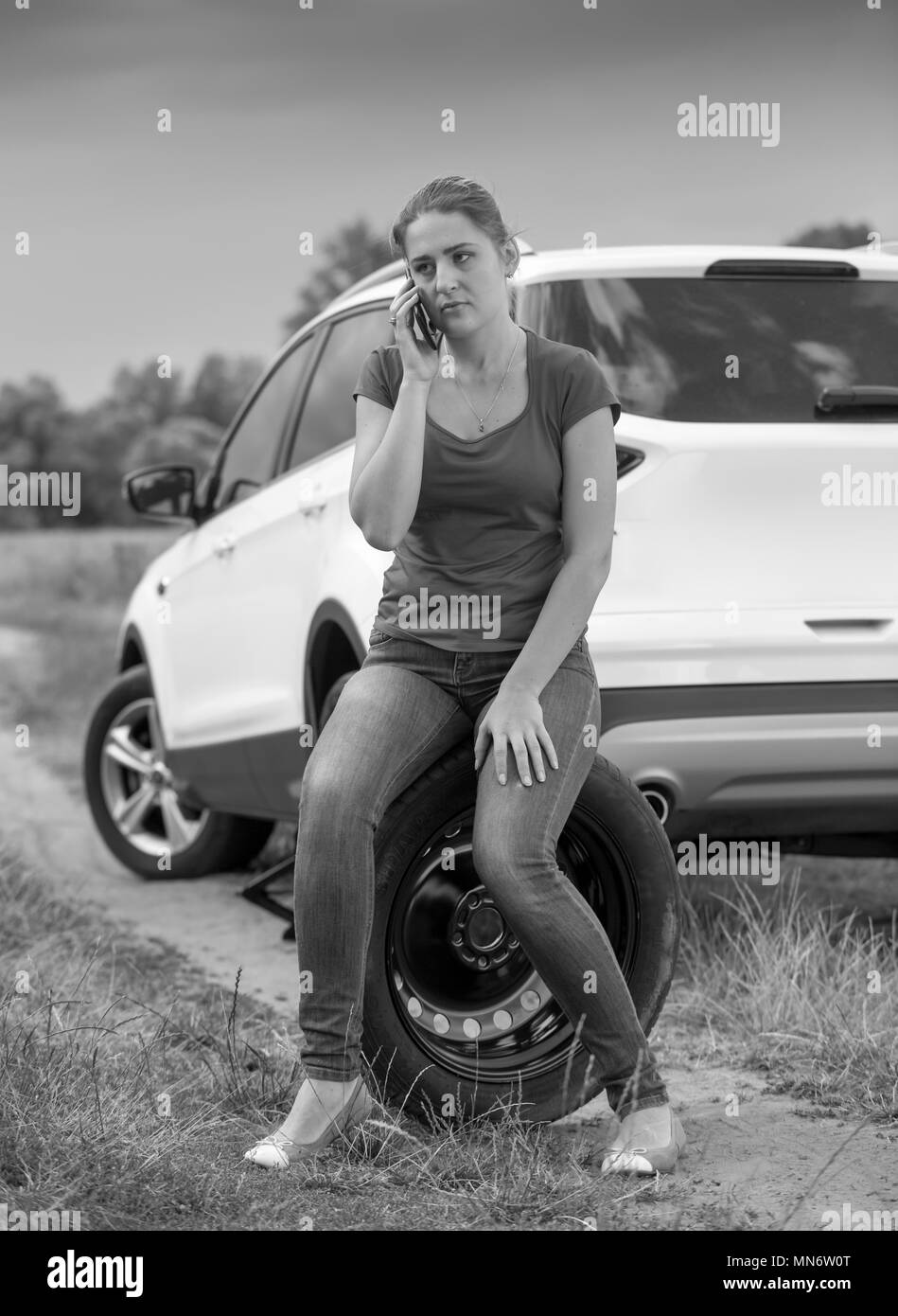 Image en noir et blanc de la jeune femme assise sur la roue de secours de voiture et d'appel dans le champ service auto Banque D'Images