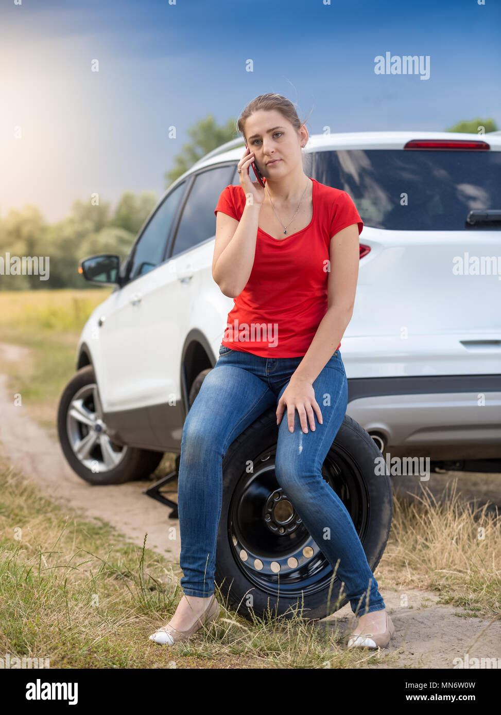 Triste jeune femme assise à côté de voiture avec roue brisée appelant auto service Banque D'Images