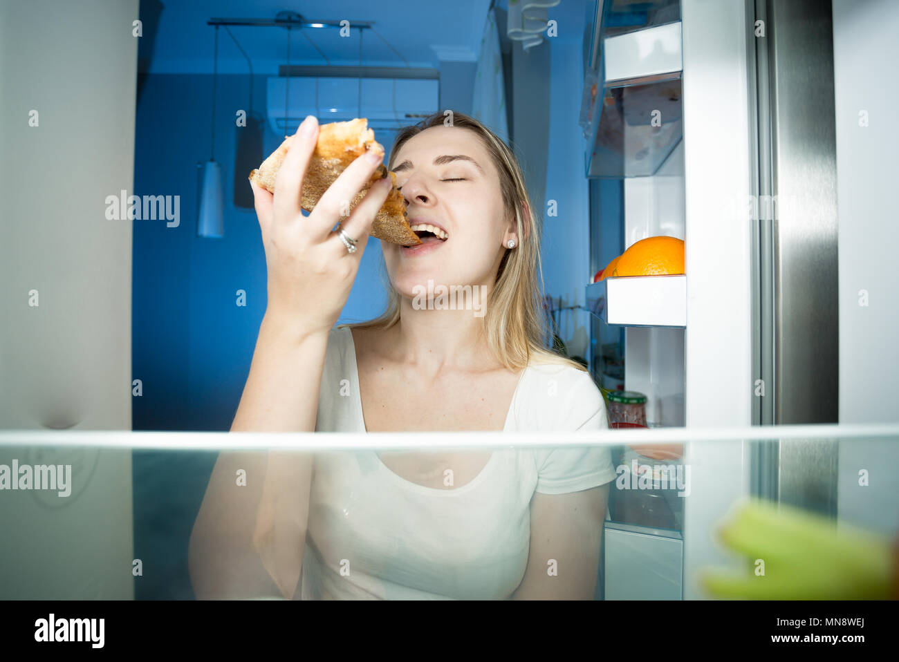 Portrait of young woman standing faim ouverte au réfrigérateur pendant la nuit et eating pizza Banque D'Images