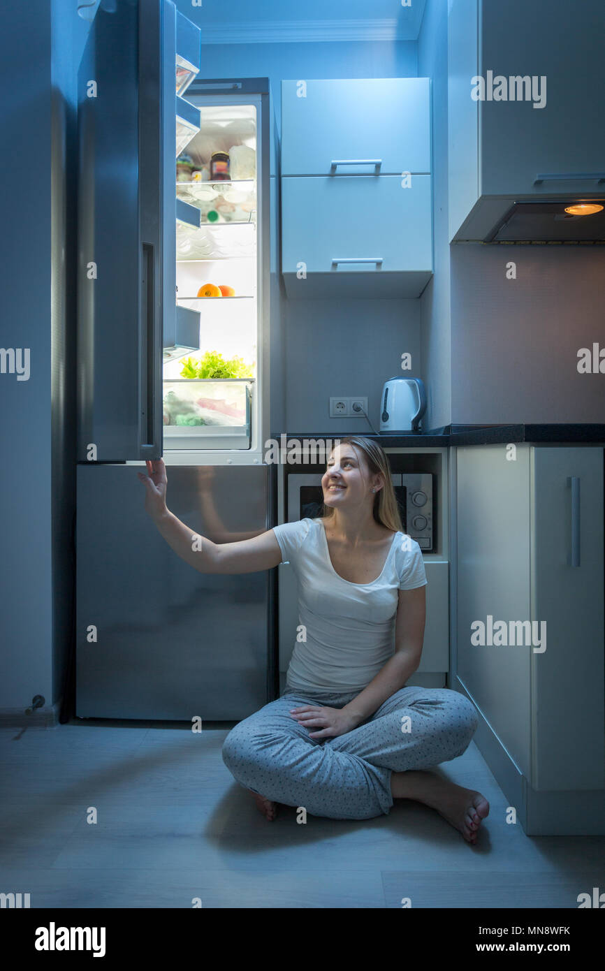 Smiling young woman sitting on cuisine dans la nuit et l'ouverture de porte du réfrigérateur Banque D'Images