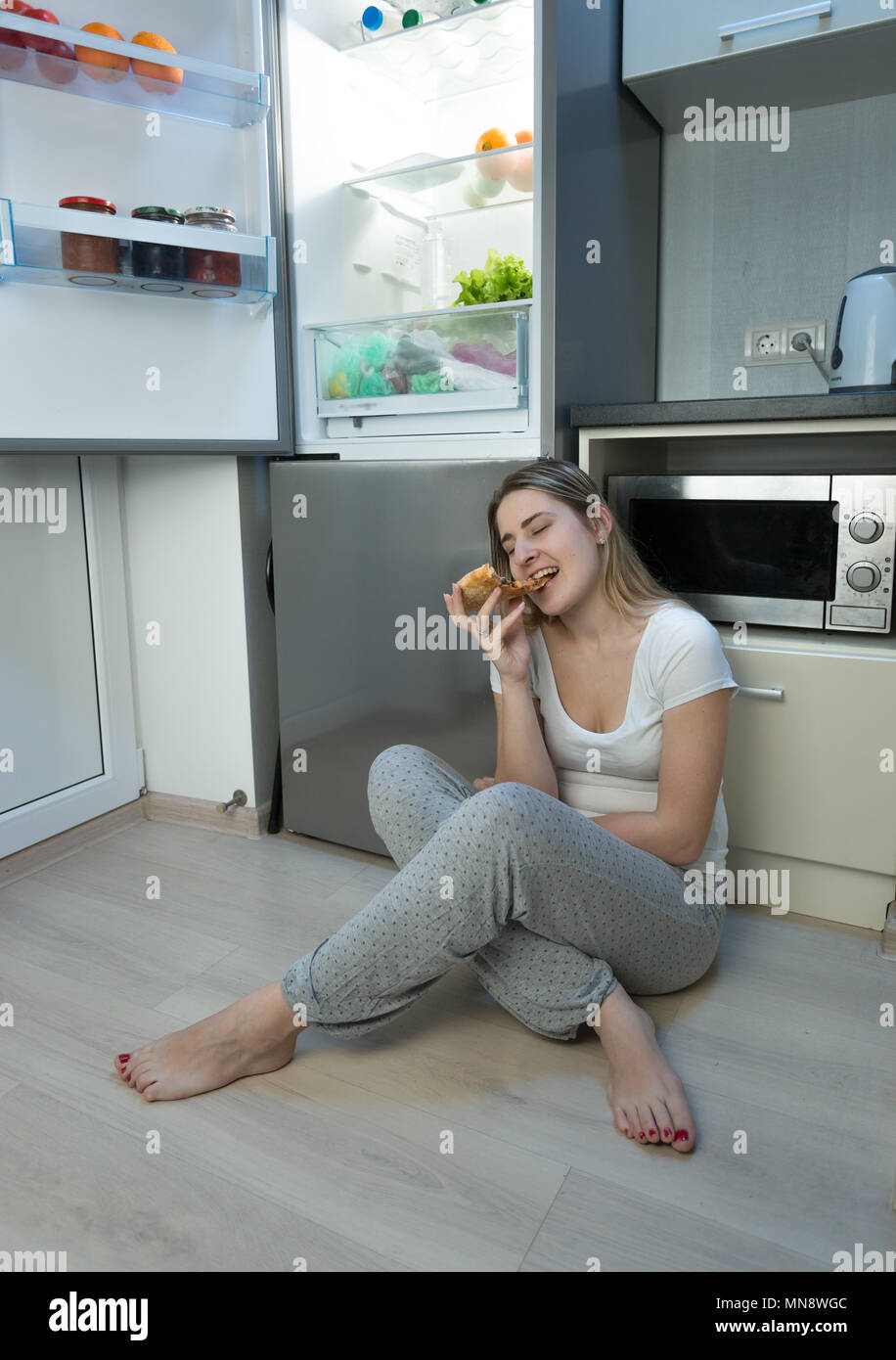 Jeune femme en pyjama assis sur le plancher de la cuisine la nuit et profiter de eating pizza Banque D'Images