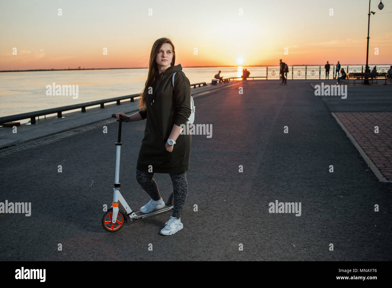 Style urbain et mode concept. Portrait d'une belle jeune femme élégante européenne modèle avec un scooter de coup sur l'asphalte sur le coucher du soleil. Banque D'Images