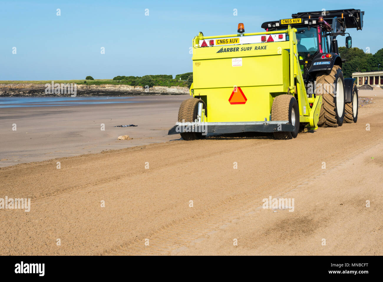 Whitmore Bay Beach à l'île de Barry, au Pays de Galles, tôt un matin d'été ensoleillé lumineux en cours de nettoyage de la litière par un tracteur rouge tirant une coiffure Surf Rake. Banque D'Images