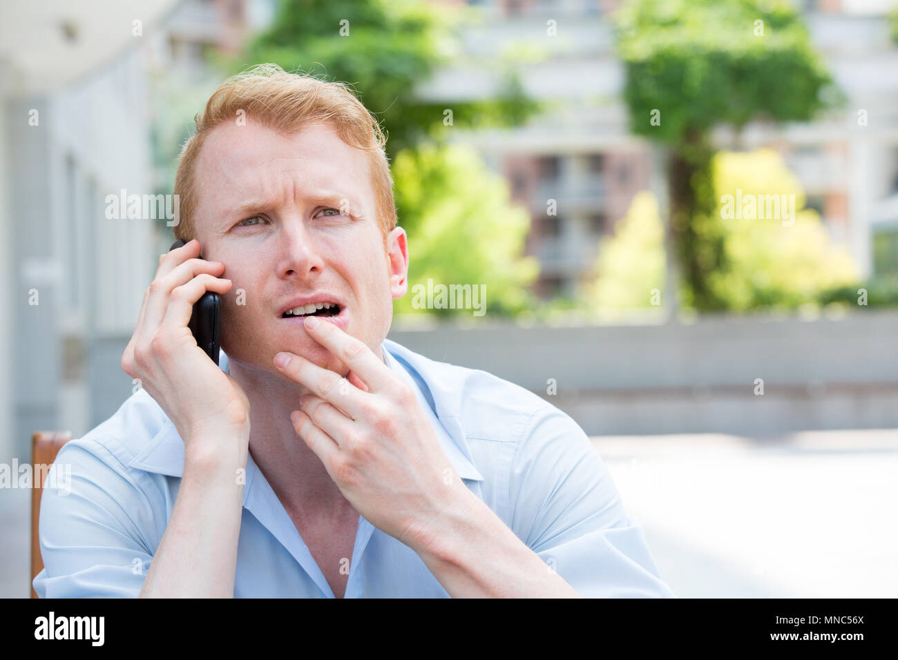 Closeup portrait, jeune homme perplexe, déconcerté, déconcerté, ahuri par quelqu'un parler sur son téléphone portable, isolé à l'extérieur contexte Banque D'Images