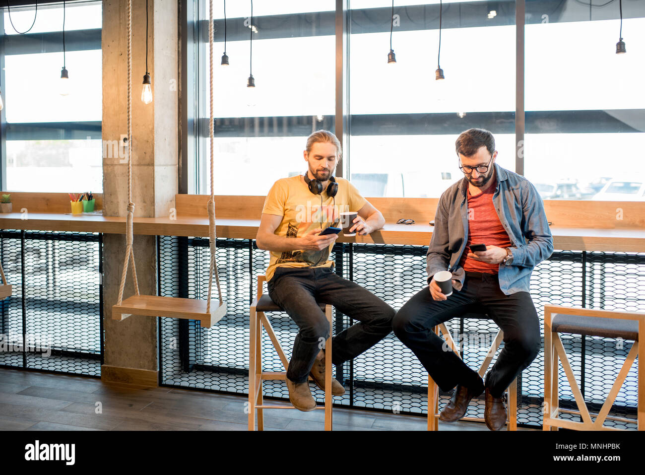 Deux hommes habillés utiliser négligemment assis smartphones près de la fenêtre de l'intérieur moderne cafe Banque D'Images
