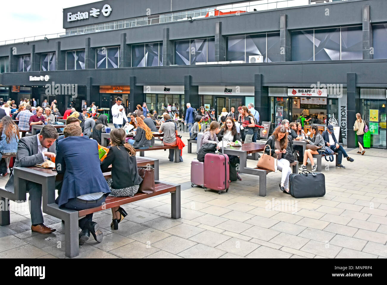Foule de gens les voyages en train d'attendre au table de pique-nique à l'extérieur de Londres Euston gare certains avec valise assurance restauration rapide café boutiques au-delà UK Banque D'Images
