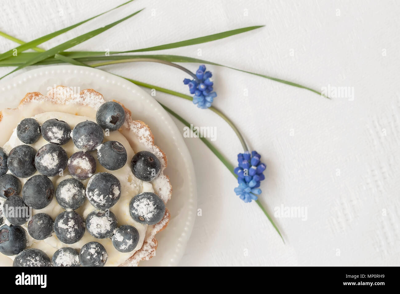 Tartelette aux bleuets frais avec crème vanille dans plat close-up, fleurs bleu, blanc serviette. De l'été romantique et délicieux dessert berry, copiez l'espace. Selective focus Banque D'Images
