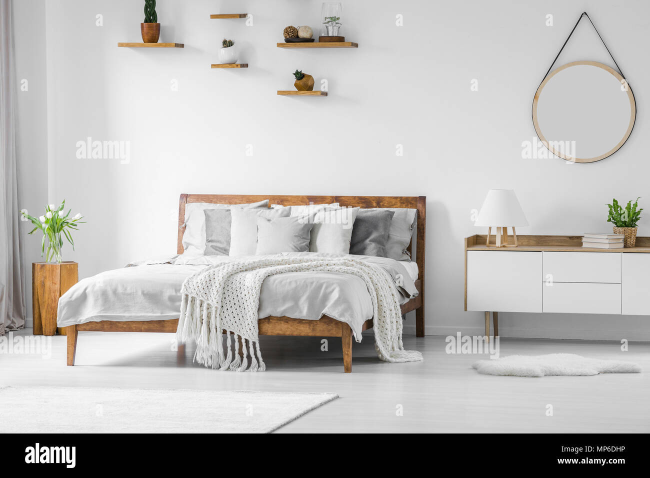 Confortable, grand lit en bois, avec des draps, oreillers et couvertures,  table à côté et miroir rond accroché sur un mur blanc dans une chambre  lumineuse de l'intérieur Photo Stock - Alamy