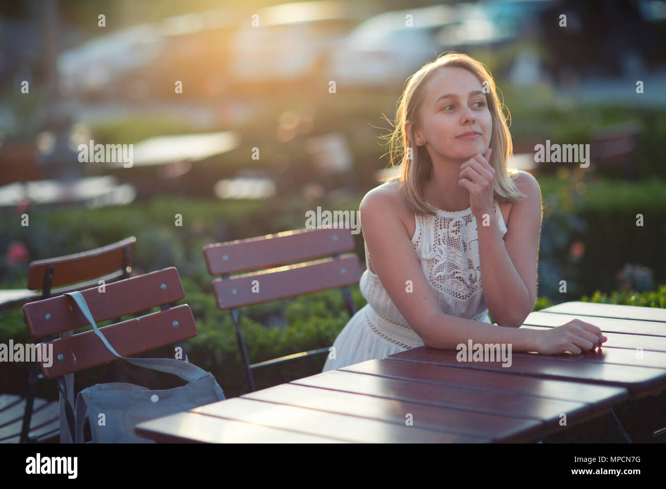 Une charmante jeune femme pensif assis à la table en bois dans le patio extérieur derrière le soleil couchant. Banque D'Images