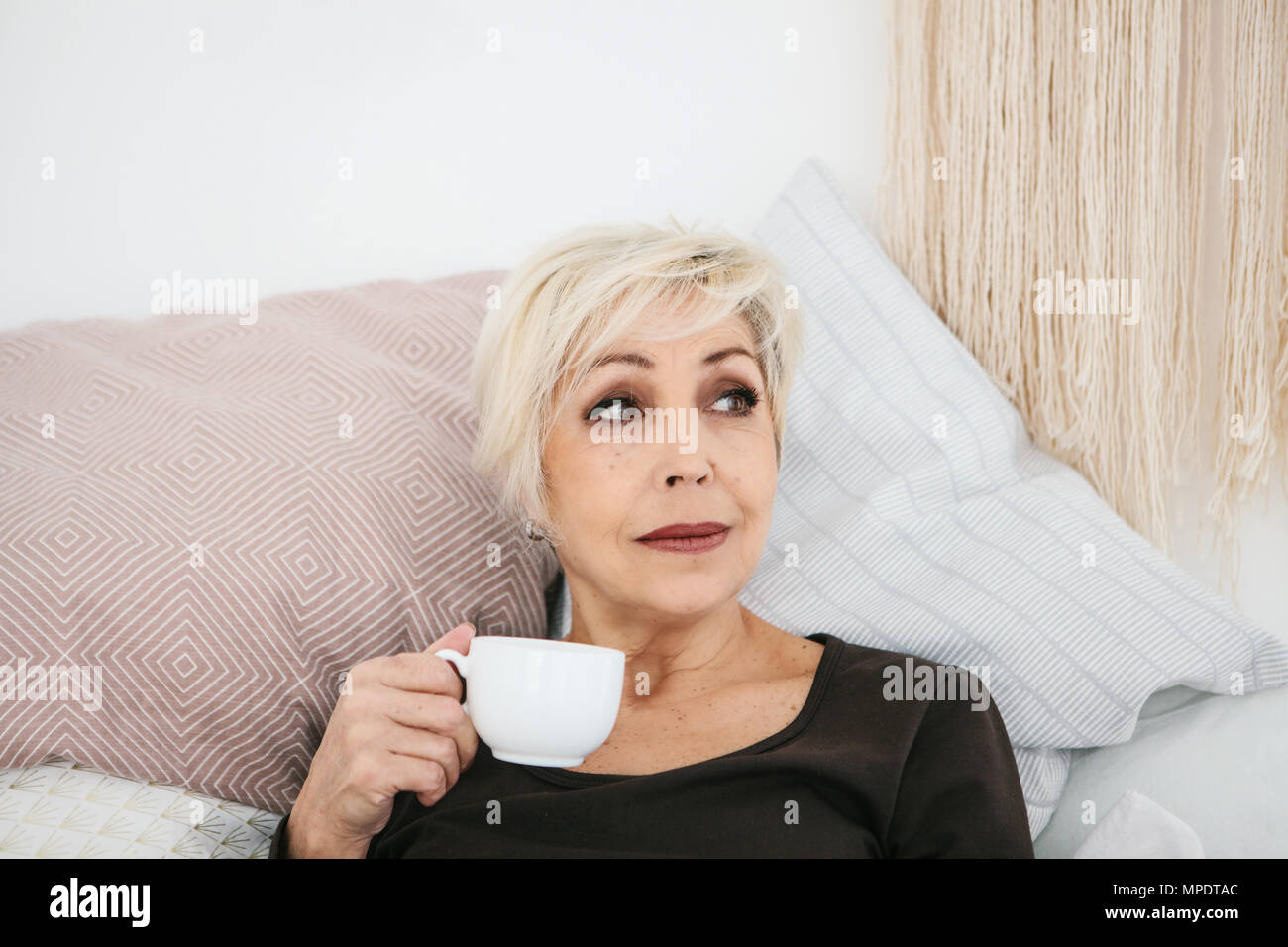 Une vieille femme solitaire se trouve au lit et boit un verre dans un mug Banque D'Images