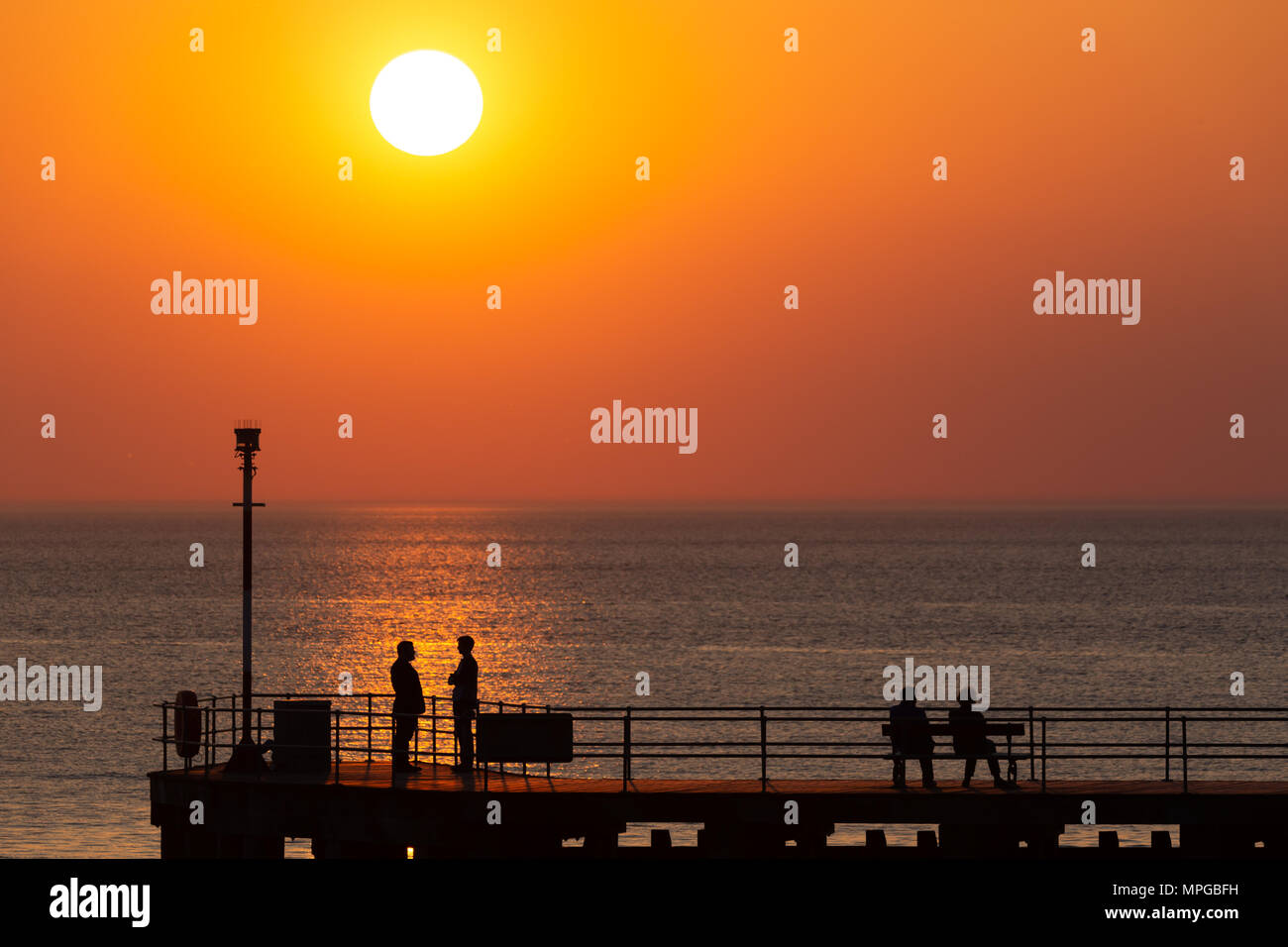 La silhouette de deux personnes assis sur une jetée en regardant le coucher du soleil sur la côte ouest du pays de Galles, tandis que deux autres se tiennent en face de l'autre parler. © Ian Jones/Alamy Live News. Banque D'Images