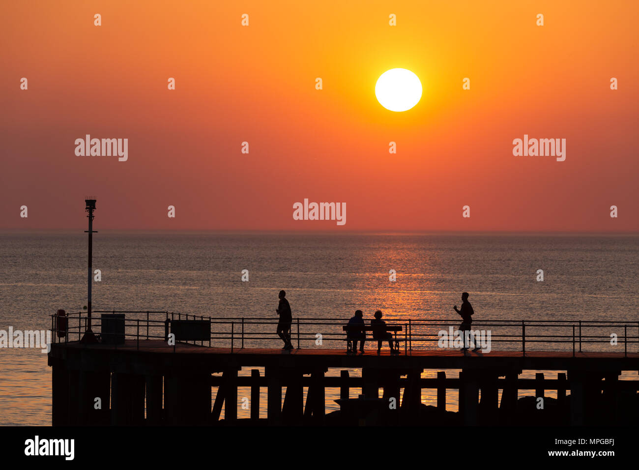 La silhouette de deux personnes assis sur une jetée en regardant le coucher du soleil sur la côte ouest du pays de Galles, tandis que deux autres jog chaque côté. © Ian Jones/Alamy Live News. Banque D'Images