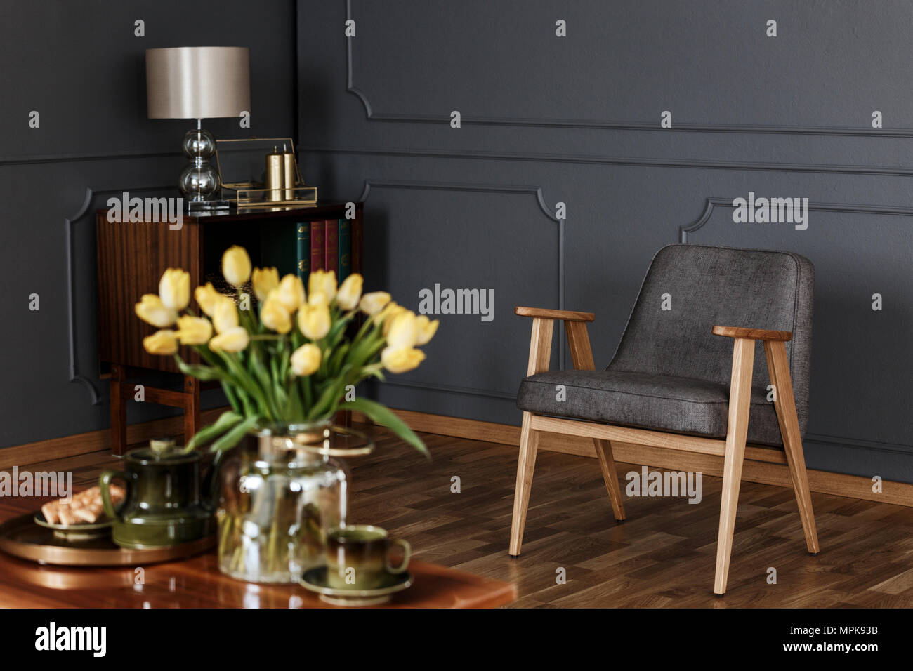 Photo réelle d'un fauteuil gris rétro et armoire en bois contre un mur sombre de la vie intérieur. Tulipes jaune floue au premier plan. Banque D'Images