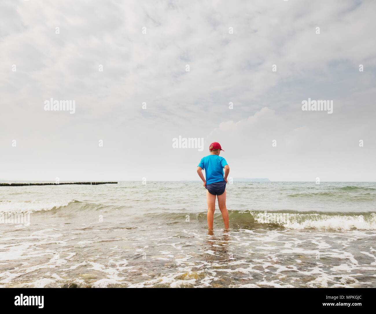 Garçon en bleu noir vêtements sportive va dans l'eau froide de la mer. Jour de vent avec la mer ondulée mousseuse. Cheveux blonds kid sur Stony Beach. Banque D'Images