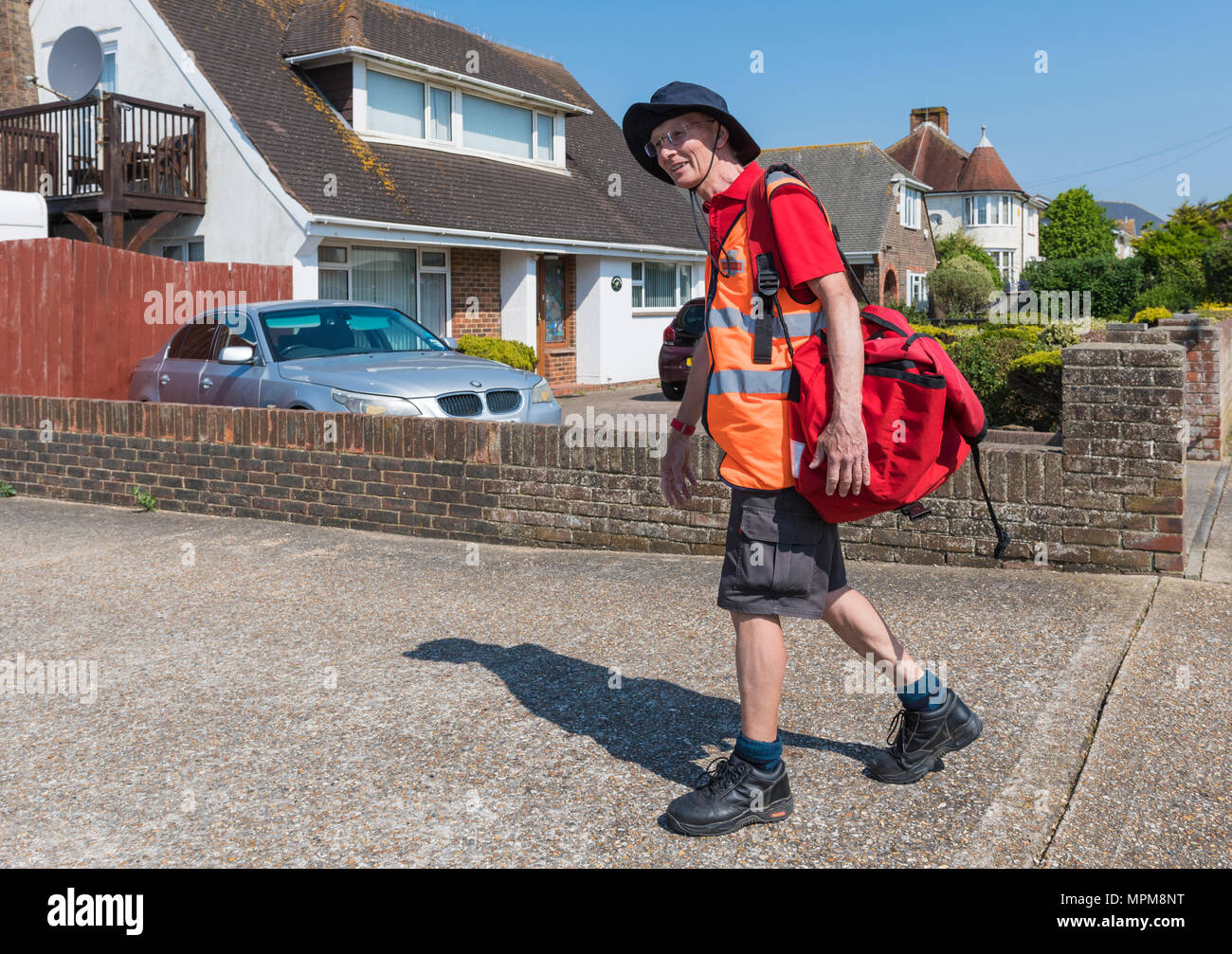 Facteur britannique portant des shorts de marcher dans une zone résidentielle portant un sac rouge de lettres, pour les livraisons du matin dans le West Sussex, Angleterre, Royaume-Uni. Banque D'Images