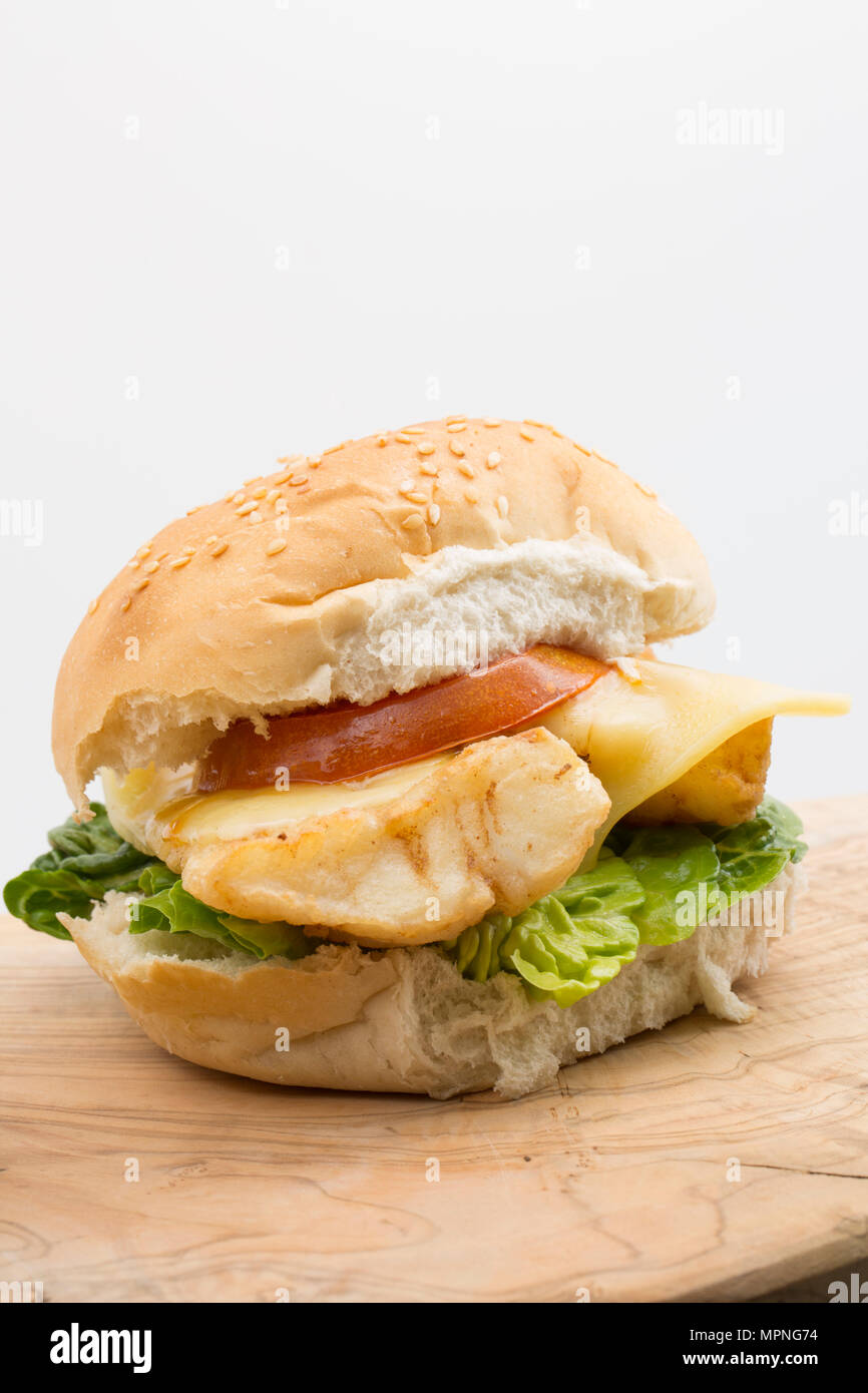 Un filet de lieu jaune, Pollachius pollachius, qui a été enduit de farine et frits, servis sur un hamburger bun avec laitue, tomates grillées et fondre Banque D'Images