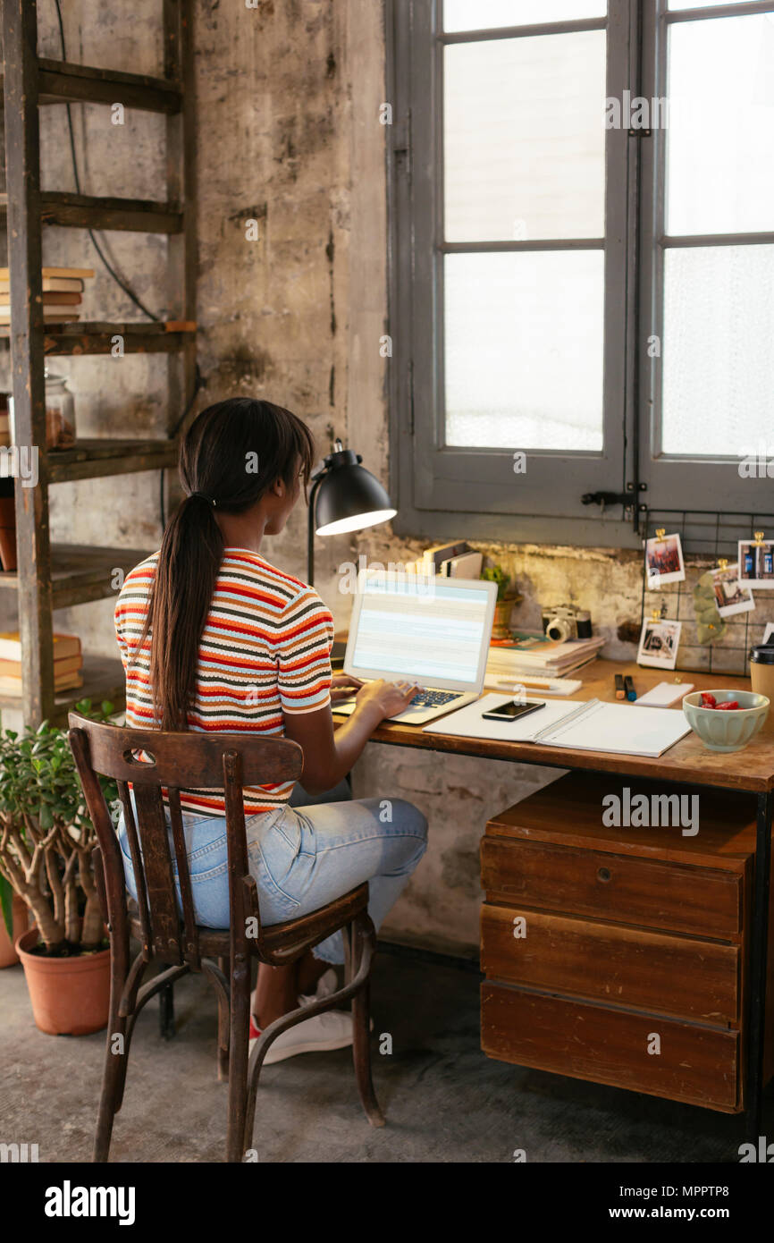 Vue arrière de young woman sitting at desk dans un loft working on laptop Banque D'Images