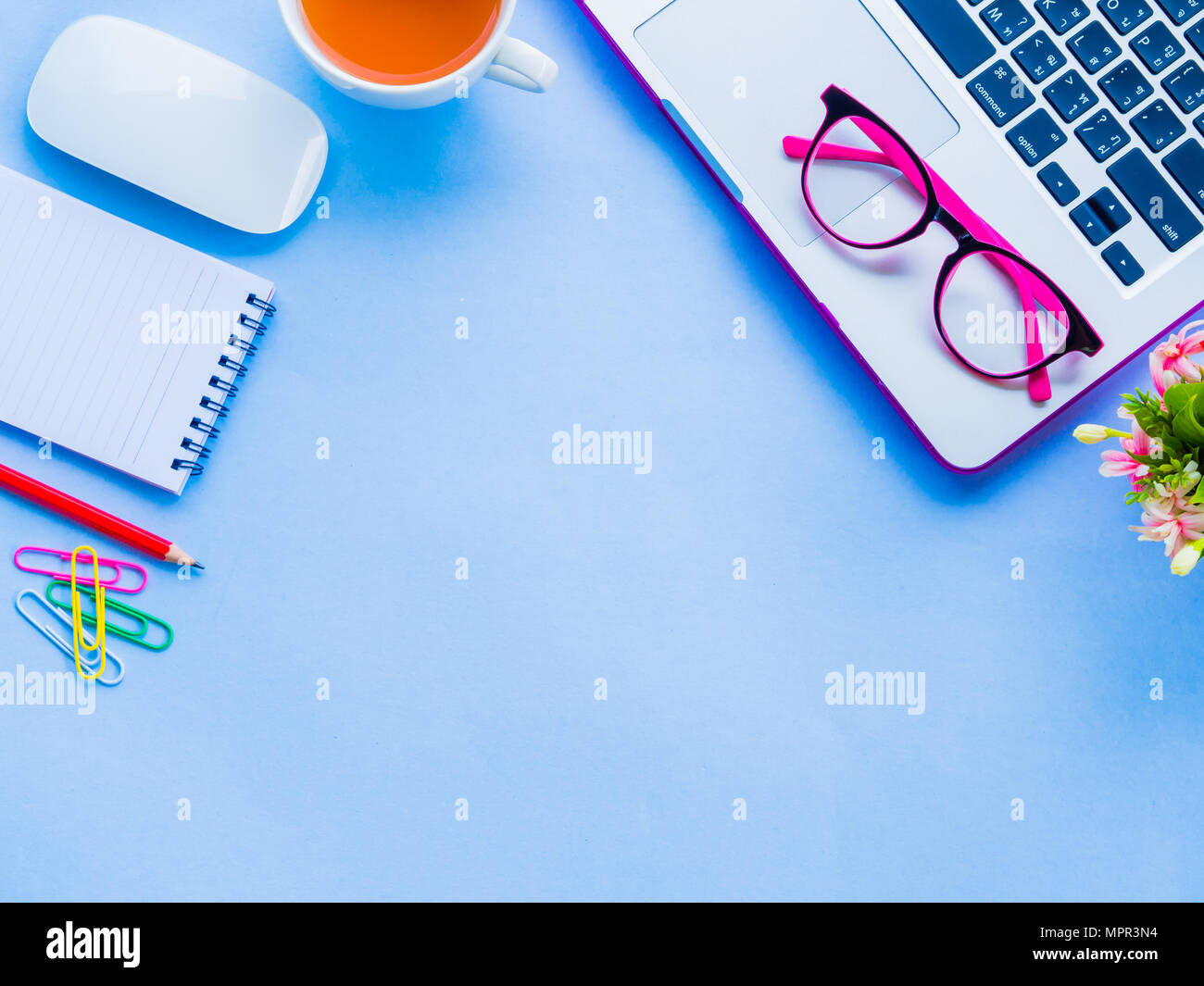 Vue de dessus de l'espace de bureau féminin avec accessoires de bureau avec ordinateur portable, carnet de notes, crayon rouge, café, fleurs, souris et lunettes roses sur bleu backg Banque D'Images