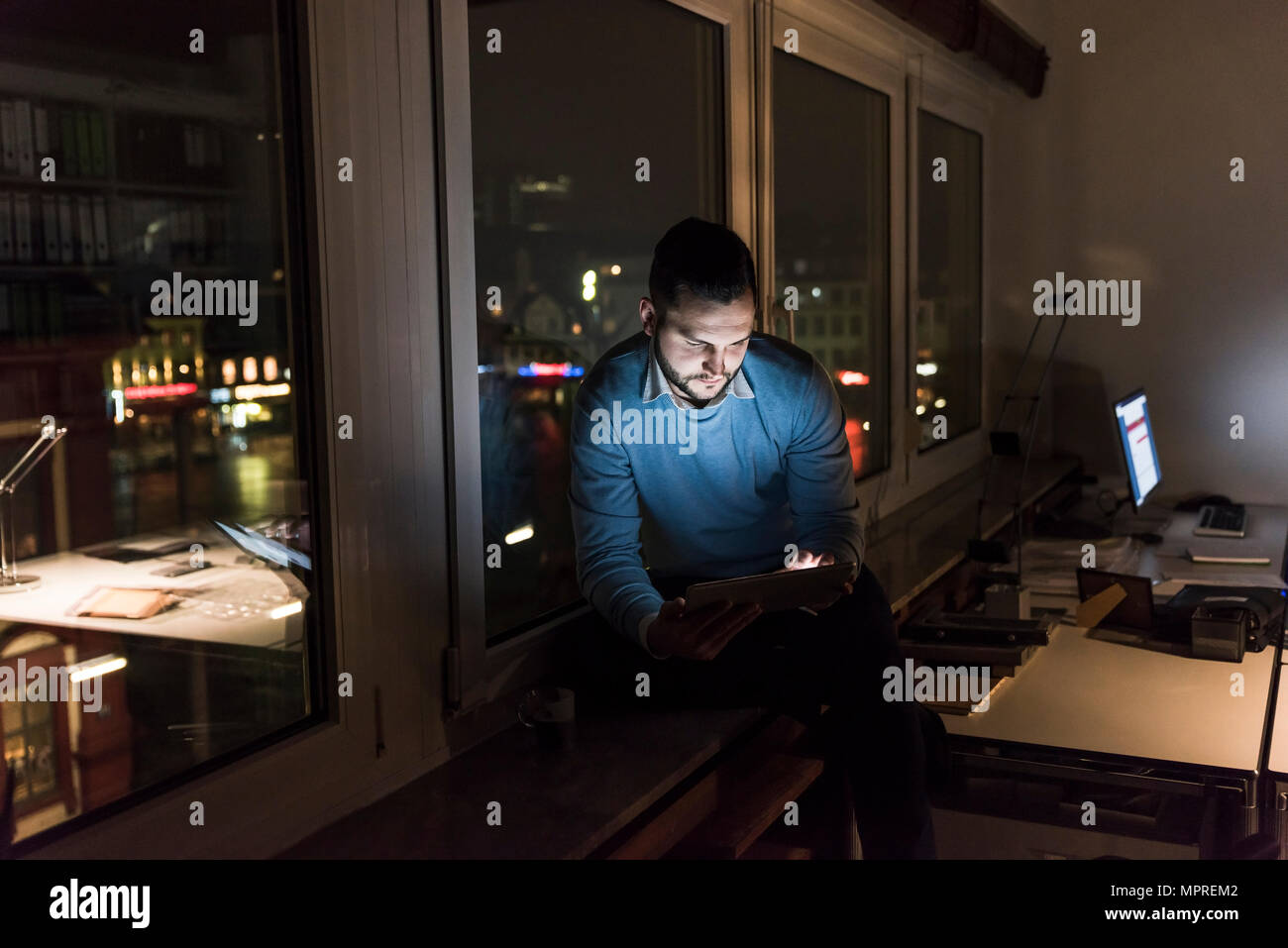 Businessman sitting on window sill dans office, la nuit, un comprimé Banque D'Images