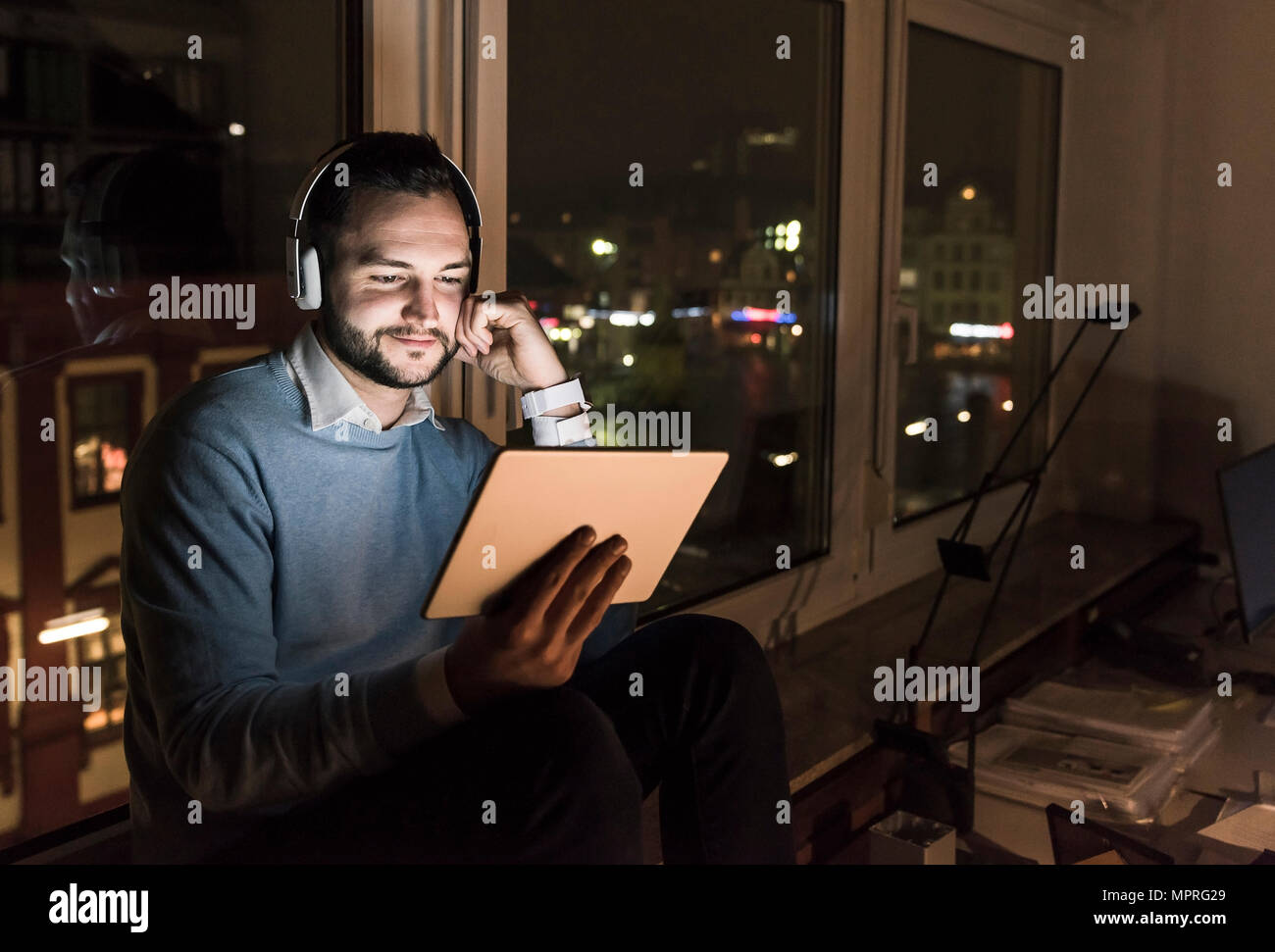 Businessman sitting on window sill dans office, la nuit, un casque et tablette Banque D'Images